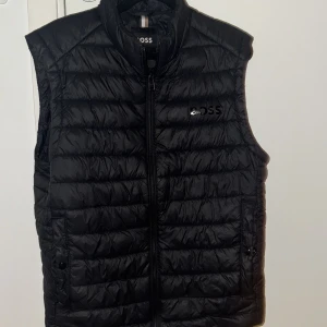 Svart dunväst från Boss - Snygg svart dunväst från Boss. Västen snålt änvänd fast fortfarande en lite fläck som sitter innanför västen( borde gå bort i tvätten). Annars e den rätt fräsch väldigt tunn och luftigt så perfekt med en hoodie under 💯🍾 (köptes i affär för 1500)