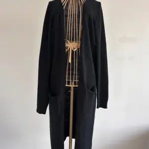 Säljer en stilren svart lång cardigan från VILA. Den har långa ärmar och praktiska fickor framtill. Perfekt för lite kyligare dagar. Enkel design som passar till många tillfällen. Stl M. Lite nopprig.