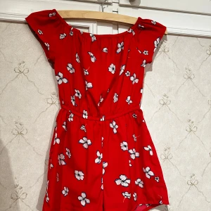 Röd blommig klänning från New Look - Söt röd byxdress (shorts och blus i samma plagg) med blommigt mönster från New Look. Klänningen har korta ärmar och en elastisk midja för en bekväm passform. Perfekt för varma dagar! STORLEK=140cl