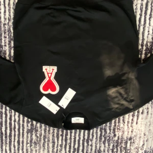 Svart tröja från Ami - Snygg svart tröja från Ami med ett stort rött hjärta och bokstaven 'A' broderat på framsidan. Tröjan har långa ärmar och en rund halsringning. Perfekt för en stilren och modern look.