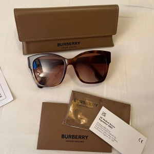 Bruna solglasögon från Burberry - Snygga solglasögon från Burberry med en brun, rektangulär båge och mörka glas. Bågarna har ett elegant Burberry-emblem på sidan. Mycket fint skick. Nypris 2098, säljer för 1000:- 