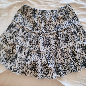 Mönstrad kjol från Lindex - Säljer en snygg mönstrad kjol från Lindex i svart och vit. Kjolen har volanger och ett elastiskt midjeband för en bekväm passform. Perfekt för att ge en trendig touch till din outfit!