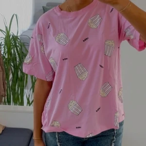Rosa t-shirt - Säljer en söt rosa t-shirt med ett roligt popcornmönster. T-shirten har en avslappnad passform och korta ärmar, perfekt för en casual look. Perfekt för filmkvällar eller bara för att liva upp vardagen! 🍿
