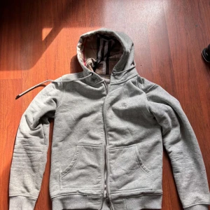 Grå hoodie från Burberry - Säljer en stilren grå hoodie från Burberry med dragkedja och klassiskt rutigt mönster i luvan. Perfekt för en avslappnad look med praktiska fickor framtill. Passar bra till både jeans och joggers. Inte fast pris kan gå lägre 