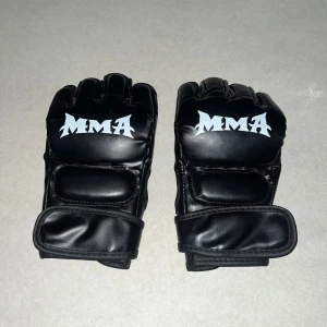 Svarta MMA-handskar - Säljer ett par svarta MMA-handskar med vit text. De är tillverkade i skinn och har öppna fingrar för bättre grepp och rörelsefrihet. Perfekta för träning och tävling. Justerbara kardborreband för optimal passform. Mått: 22cm längd och 12 cm bredd.