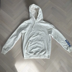 Vit hoodie från GAP - Säljer en vit hoodie från GAP med blå och gul logga. Den har en stor ficka framtill och justerbar huva med snören. Perfekt för en avslappnad stil.