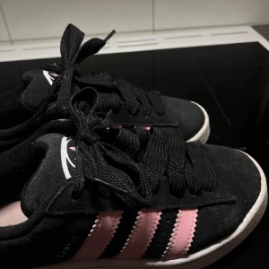 Svarta och rosa sneakers från Adidas campus  - Snygga svarta sneakers från Adidas med rosa ränder på sidorna. Skorna har en klassisk design med snörning och en vit sula som ger en stilren. De är använda förut.