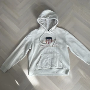 Vit hoodie från GANT - Säljer en stilren vit hoodie från GANT med en broderad logotyp och känguruficka. Perfekt för en avslappnad stil. Har en liten fläck på vänster ärm, men inget märkbart. Huvan har dragsko och tröjan är tillverkad i mjukt material. Storlek L, skulle även tro att den passar M. 