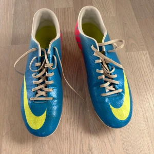 Blå Nike Mercurial fotbollsskor - Säljer ett par blå Nike Mercurial fotbollsskor med gula och rosa detaljer. Skorna har snörning och är designade för optimal prestanda på planen. Perfekta för fotbollsspelare som vill ha stil och funktionalitet. Storlek 44.5. Sparsamt använda sista bilden ser man de börjat släppa lite.