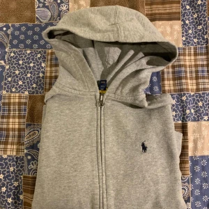 Grå hoodie med huva från Polo Ralph Lauren - Säljer en grå hoodie med huva från Polo Ralph Lauren. Hoodien har en dragkedja framtill och är dekorerad med den klassiska logotypen på bröstet. Perfekt för en avslappnad stil. Passar åldern 10-12