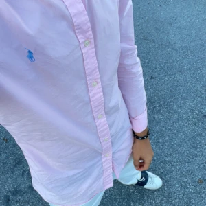 Rosa skjorta från Ralph Lauren - Snygg rosa skjorta från Ralph Lauren med klassisk krage och knappar framtill. Skjortan har en broderad logotyp i blått på bröstet och långa ärmar med knappslå vid ärmsluten. Perfekt för en stilren look.