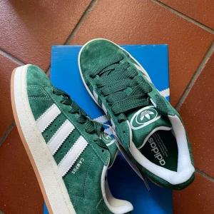 Gröna Adidas Campus sneakers - Snygga gröna Adidas Campus sneakers i mocka med klassiska vita ränder och snörning. Perfekta för en stilren och sportig look. Gummisula i beige ger en skön komfort och bra grepp. Använda max 3-4 gånger (se på sulan)