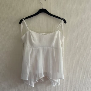 Vit ärmlös topp från Nelly - Säljer en söt vit ärmlös topp från Nelly med tunna axelband och volangdetaljer. Toppen har en luftig och fladdrig design, perfekt för varma dagar. Den har en smockad rygg för extra komfort och stil. Passar perfekt till sommarens alla tillfällen! 🌸Helt oanvänd