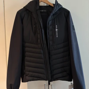 Svart hybridjacka från Sail Racing - Snygg svart hybridjacka från Sail Racing med dragkedja och huva. Jackan har en modern design med quiltade detaljer på sidorna och en logotyp på bröstet. Perfekt för kyliga dagar.