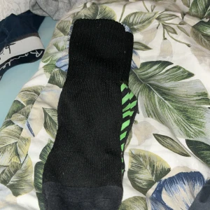 Svarta strumpor med gröna detaljer - Säljer ett par svarta strumpor med gröna halkskyddsdetaljer på undersidan. Strumporna är ifrån unisports hemsida. Perfekta för att hålla fötterna varma och säkra på hala ytor.