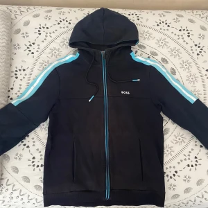 Svart hoodie från Boss med blå detaljer - Snygg svart hoodie från Boss med dragkedja och huva. Den har blå ränder längs ärmarna och blå detaljer vid dragkedjan. Perfekt för en casual look. Har endast ett fel i en av trådarna annars är den helt felfri förutom armen då också som är köpt för 2400kr från thernlunds