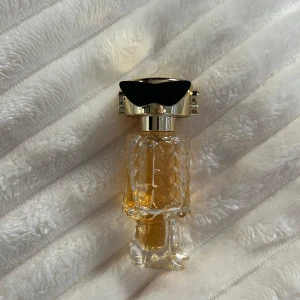 Paco rabanne fame  - Elegant parfymflaska med en unik design som påminner om fjäll. Flaskan har en gyllene vätska och ett lock i glänsande guld och svart. Perfekt för den som vill ha en exklusiv touch på sin doftsamling.