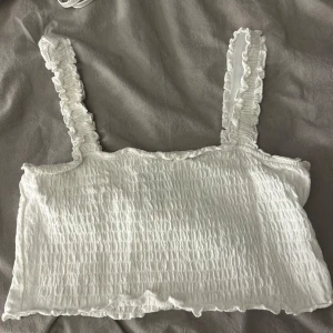 Vit ribbad linnetopp från Vero Moda - Säljer en söt vit ribbad linnetopp från Vero Moda. Toppen har volangdetaljer på axelbanden och en smockad textur som ger en fin passform. Perfekt för varma sommardagar eller som ett lager under en jacka.💞