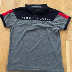 Grå pikétröja från Tommy Hilfiger - Snygg grå pikétröja från Tommy Hilfiger med svarta och röda detaljer på axlarna. Tröjan har en klassisk krage och korta ärmar. Perfekt för en stilren och avslappnad look.