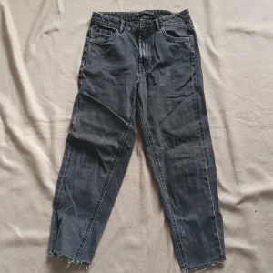 Grå jeansbyxor med råa benslut - Säljer ett par grå jeansbyxor från Zara med raka ben och råa, avklippta benslut. Klassisk femficksmodell med bälteshällor och knappgylf. Perfekta för en avslappnad och trendig look. Cropped längd 
