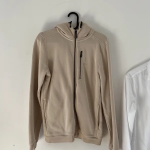 Beige sail racing hoodie med dragkedja - Säljer en stilren beige sail racing hoodie med dragkedja framtill och huva. Tröjan har en bröstficka med dragkedja och två sidofickor. Perfekt för en avslappnad look och enkel att matcha med det mesta.