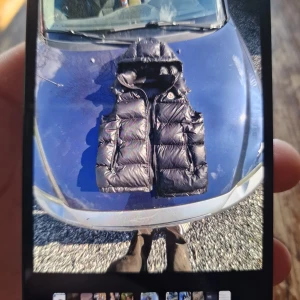 Svart dunväst med huva - Säljer en svart dunväst med huva och dragkedja framtill. Västen har en glansig yta och är vadderad för extra värme. Perfekt för lager-på-lager under kyliga dagar.