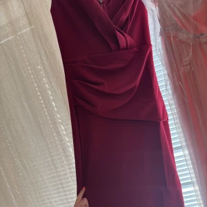 Vinröd långklänning från Ax Paris - Säljer en elegant vinröd långklänning från Bubbleroom med draperad detalj framtill och omlott vid bysten. Klänningen har en figurnära passform och är perfekt för festliga tillfällen.