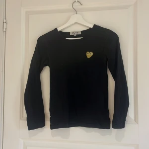 Svart långärmad tröja från Comme des Garçons Play - Svart långärmad tröja från Comme des Garçons Play med det ikoniska gula hjärtat broderat på bröstet. Enkel och stilren design med rund halsringning. Perfekt för dig som gillar minimalistisk stil och vill ha något från ett känt märke.