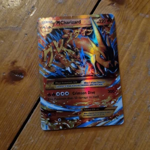 Pokémonkort M Charizard EX - Säljer ett Pokémonkort av typen M Charizard EX med holografisk yta. Kortet har färger i guld, rött, blått och svart och visar Charizard i en dynamisk pose. Text och detaljer är tryckta på engelska. Baksidan har klassiskt blått Pokémon-motiv.                       Obs upphämtning här eller i Älmhult 