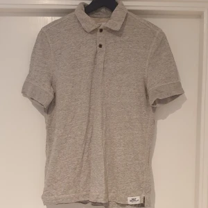 Ljusgrå pikétröja från David Beckham x H&M - Säljer en stilren ljusgrå pikétröja från David Beckham Bodywear för H&M. Tröjan har klassisk krage, två knappar och korta ärmar. Perfekt för en avslappnad men snygg look.