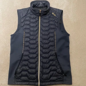 Marinblå dunväst från BOSS - Marinblå svart dunväst från BOSS med quiltad framsida och dragkedja hela vägen. Västen har hög krage och diskret BOSS-logga på bröstet. Perfekt att bära över en tröja för extra värme och stil.
