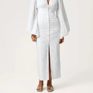 Vit långklänning med puffärmar och knappar - Elegant vit långklänning med v-ringning, markerad midja och långa puffärmar. Klänningen har dekorativa knappar framtill och ett subtilt blommigt mönster i tyget. Perfekt för sommarens speciella tillfällen.