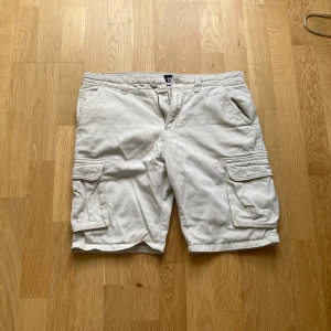 Beige cargoshorts från 157 - Säljer ett par vita cargoshorts från 157 med flera praktiska fickor på sidorna och klassisk knapp- och dragkedjestängning. Perfekta för sommardagar när du vill ha en avslappnad och funktionell stil.