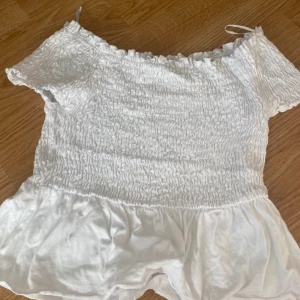 Vit offshoulder topp från Gina Tricot - Säljer en söt vit offshoulder topp från Gina Tricot. Toppen har smockad överdel och volang nertill, vilket ger en luftig och somrig känsla. Perfekt till varma dagar och enkel att matcha med jeans eller kjol.Frakt tillkommer