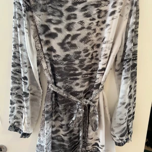 Grå leopardmönstrad klänning från Dry Lake - Säljer en snygg långärmad klänning från Dry Lake i grått och svart leopardmönster. Klänningen har ett knytband i midjan och är tillverkad i ett lätt, glansigt material som ger en elegant känsla.