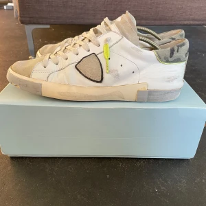 Vita sneakers från Philippe Model - Säljer ett par vita Philippe Model sneakers med grå detaljer och en gul snörningsdetalj på sidan. Skorna har en klassisk låg profil, rund tå och platt sula. Märkets logotyp syns tydligt på sidan. Perfekta för en stilren och avslappnad look. 8/10 skick priset, är inte hugget i sten!