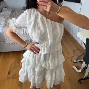 Vit oneshoulderklänning med volanger - Superfin vit klänning med oneshoulder och volanger i flera lager. Klänningen har ett avtagbart knytband i midjan. Passar perfekt till studenten🌻 Klänningen helt oanvänd och endast provad.