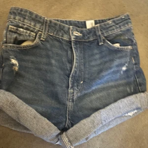 Blå jeansshorts relaxed high waist - Snygga blå jeansshorts med relaxed passform och mid waist midja. Shortsen har upprullade benslut, slitna detaljer och klassiska fem fickor. Perfekta för en avslappnad stil under varma dagar. 