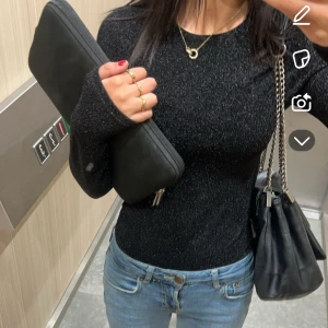 Svart glittrig långärmad topp - Säljer en svart glittrig långärmad body med tight passform. Perfekt att styla med jeans eller kjol för en snygg look. Materialet är stretchigt och skimrar snyggt i ljuset. Kedjeväska och accessoarer ingår ej.