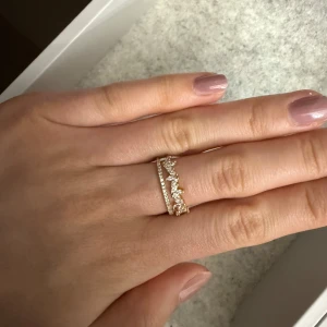 Guldfärgad ring med stenar - Säljer en snygg guldfärgad ring med glittriga stenar i en unik design. Perfekt för att ge din look lite extra bling. Ringen har en öppen baksida vilket gör den justerbar i storlek.