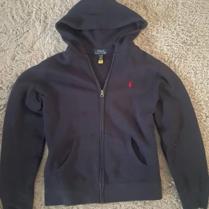 Mörkblå hoodie från Polo Ralph Lauren - Mått: bröst 50cm längd 56cm ärm 55cm Säljer en mörkblå hoodie från Polo Ralph Lauren med dragkedja och huva. Tröjan har två fickor framtill och en liten röd broderad logga på bröstet. Perfekt för en avslappnad stil.