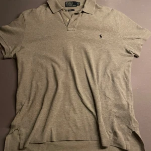 Grå pikétröja från Polo Ralph Lauren - Säljer en klassisk grå pikétröja från Polo Ralph Lauren med broderad logga på bröstet. Tröjan har korta ärmar, krage och knappar framtill. Modellen är custom fit och passar perfekt till en avslappnad stil. Observera att den sitter som M/L!!!!!