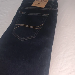 Mörkblå  slim fit jeans från Hollister - Säljer ett par mörkblå super slim fit jeans från Hollister med klassisk femficksdesign och kontrastsömmar. Byxorna har en läderpatch med logga bak i midjan och smal passform hela vägen ner.