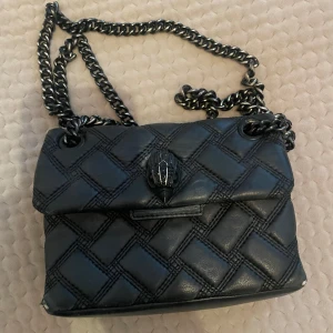 Svart quiltad axelväska med kedjerem - Säljer en svart  axelväska med kedjerem och dekorativt spänne framtill. Väskan har ett rutigt mönster i sömmarna och är tillverkad i skinnimitation. Perfekt för dig som vill ha en snygg accessoar med coola detaljer. Min väska är välanvänd och har några defekter mid märket och i hörnen av väskan, även inuti väskan i tyget💓Tryck ej på köp nu