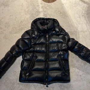 Svart dunjacka från Moncler - Säljer en svart, glansig dunjacka från Moncler med huva och dragkedja framtill. Jackan har två snedställda fickor med dragkedja och klassisk Moncler-logga på ärmen. Perfekt för kalla dagar.