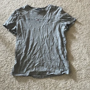 Grå t-shirt från Tommy Hilfiger - Snygg grå t-shirt från Tommy Hilfiger med tryckt logga och texten 'EST. 1985' på bröstet. Tillverkad i mjuk ekologisk bomull och har en klassisk rund halsringning. Perfekt för en avslappnad stil.