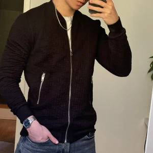 Svart overshirt  - Säljer en svart bomberjacka från J.Lindeberg med snyggt diskret mönster och silverfärgade dragkedjor. Jackan har lång ärm, ribbade muddar och klassisk bomberkrage. Perfekt för dig som gillar stilrena plagg med lite edge.