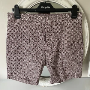 Bruna mönstrade shorts från Zara Man - Säljer ett par bruna shorts från Zara Man med ett sunikt geometriskt mönster i vitt. Shortsen har normal passform och stängs med dragkedja och knapp. Perfekta för varma dagar när du vill sticka ut lite extra.