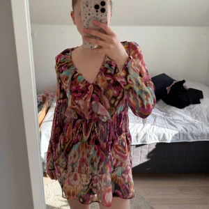 Färgglad kortklänning med volang och v-ringning - Säljer en färgglad kortklänning med långärmade transparenta ärmar och v-ringning. Klänningen har volangdetaljer framtill och ett smockat midjeparti. Mönstret är livligt i rosa, rött, blått och beige. Perfekt för dig som gillar färg och mönster! Klänning är i kortaste laget för mig som är 172 så rekommenderas för någon som är lite kortare