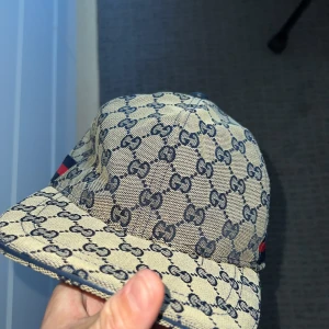 Navy keps från Gucci - Säljer en beige keps från Gucci med blått GG-mönster och en röd-blå rand på sidan. Kepsen har böjd skärm och är tillverkad i bomullsmaterial. Perfekt för dig som vill ha en snygg och lyxig accessoar. Kommer med original påsen! Storlek M.